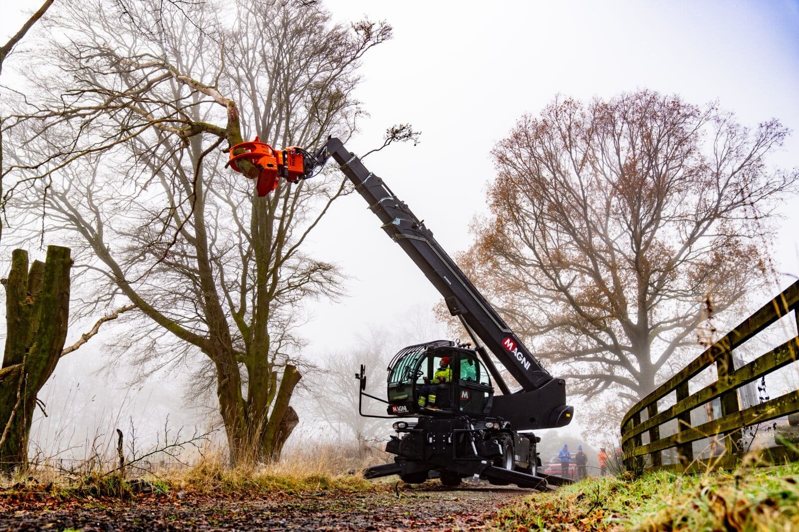 Telehandler trimming high tree branches with cutting attachment in misty countryside
