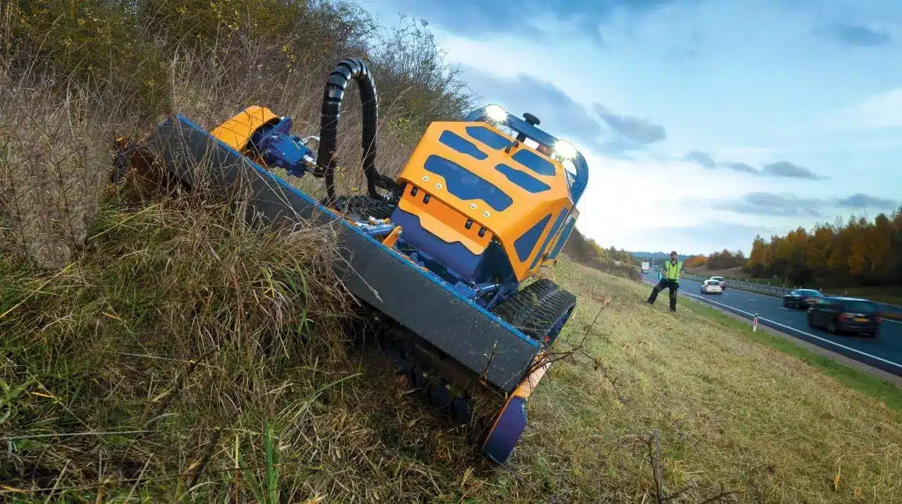 A Bomford Turner Remote‑controlled Flailbot mower cutting grass on a steep roadside embankment beside a highway.