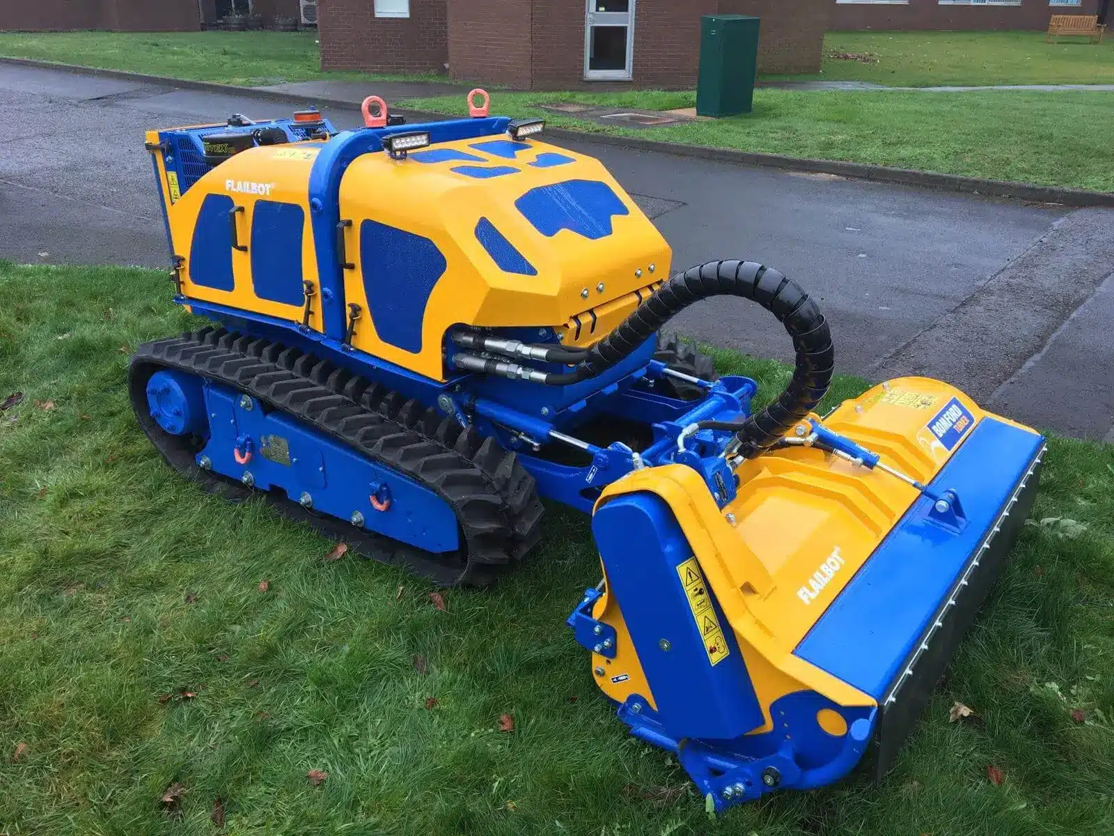 Bomford Flailbot Excel remote‑controlled tracked mower on grass near buildings.